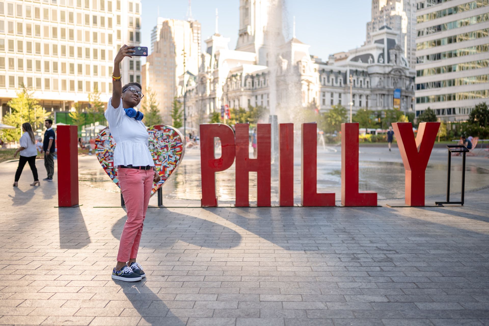 A person in white shirt and red pants taking a selfie in front of a red I "heart" philly sign