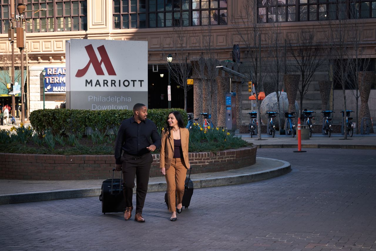 A couple walk with their luggage in front of a Marriott sign with a brick and stone hotel in the background
