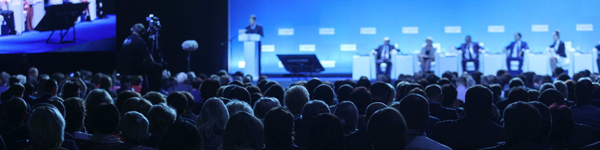 Large blue video screen backdrop behind five panelists sitting on a stage. Lots of silhouettes of the backs of audience members in the foreground.