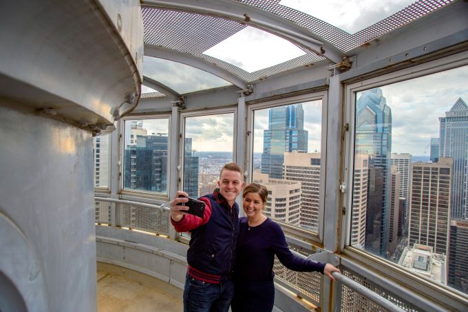 A couple at the top of the city hall tower takes a selfie in front of large glass windows with the Philadelphia city skyline behind them