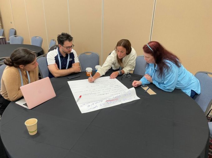 A group of four people sit at a round table writing notes on a giant paper pad during a conference breakout session